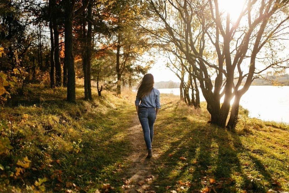 Mujer caminando por un sendero junto a un lago entre árboles iluminados por el sol